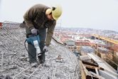 A person wearing safety equipment uses a demolition hammer to break concrete on a rooftop.