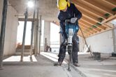 A person wearing safety equipment uses a demolition hammer to break a concrete floor.