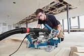 A person wearing safety equipment uses a sliding mitre saw to cut wooden planks.