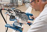 A person wearing safety equipment cuts wood with a sliding mitre saw in a workshop.