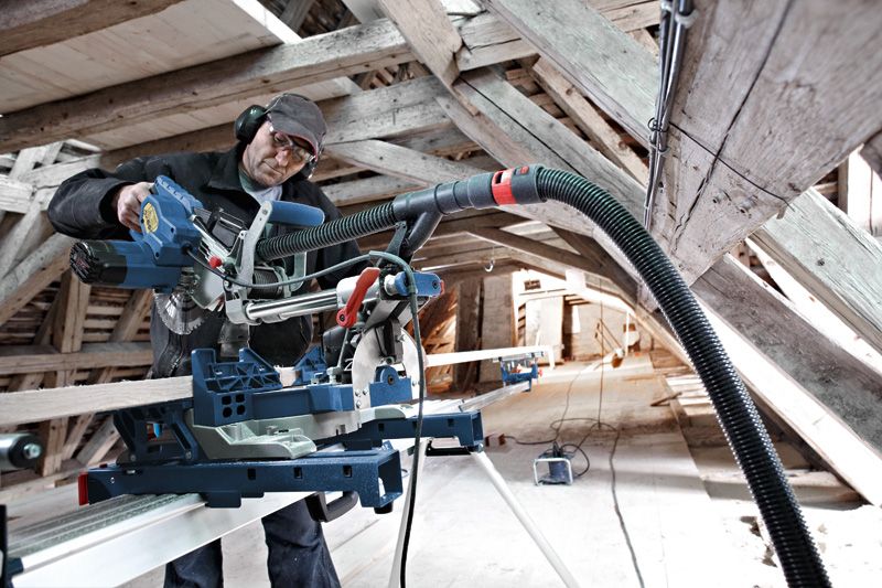 A person wearing safety equipment operates a sliding mitre saw to cut wood in an attic.