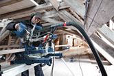 A person wearing safety equipment operates a sliding mitre saw to cut wood in an attic.