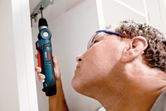 A person wearing safety equipment uses a cordless angle drill to work inside a cabinet.