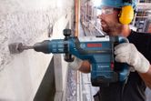 A person wearing safety equipment uses a demolition hammer to break concrete wall.