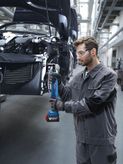 A person wearing safety equipment tightens bolts on a car frame using a cordless power tool.