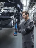 A person wearing safety equipment tightens bolts on a car frame using a cordless power tool.