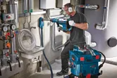A person wearing safety equipment drills into a wall near pipes using a rotary hammer.