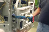 Person wearing safety equipment uses a cordless tool to fasten a hinge inside a car door.