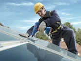 A person wearing safety equipment tightens a bolt on a rooftop solar panel with a power tool.