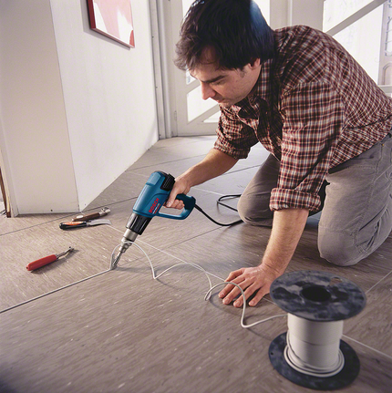 A worker kneels to heat shrink tubing onto a wire with a heat gun on a tiled floor.