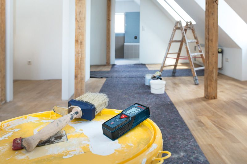 Laser measure and paintbrush rest on a bucket in a renovated attic with a ladder.