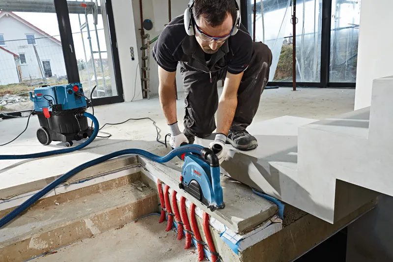 A person wearing safety equipment uses a wall chaser on concrete above exposed pipes.