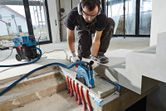 A person wearing safety equipment uses a wall chaser on concrete above exposed pipes.