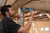 Person checks a wooden beam angle with a digital inclinometer under a timber roof.