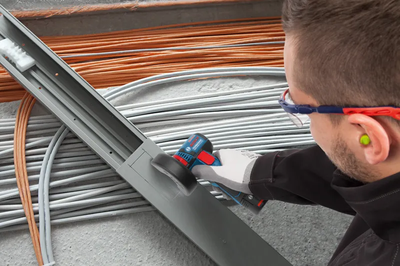 GWS 12V-76 A person wearing safety equipment cuts a metal cable tray with a cordless angle grinder.