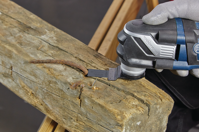 A worker wearing safety equipment uses a multi-tool to cut a rusty nail from wood.