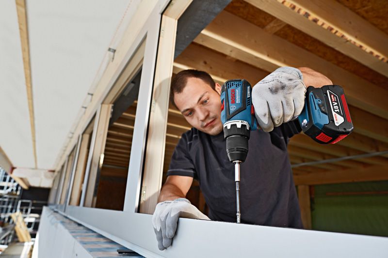 A person wearing safety equipment uses a cordless drill to drive a screw into a window frame.