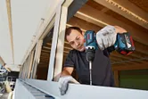 A person wearing safety equipment uses a cordless drill to drive a screw into a window frame.