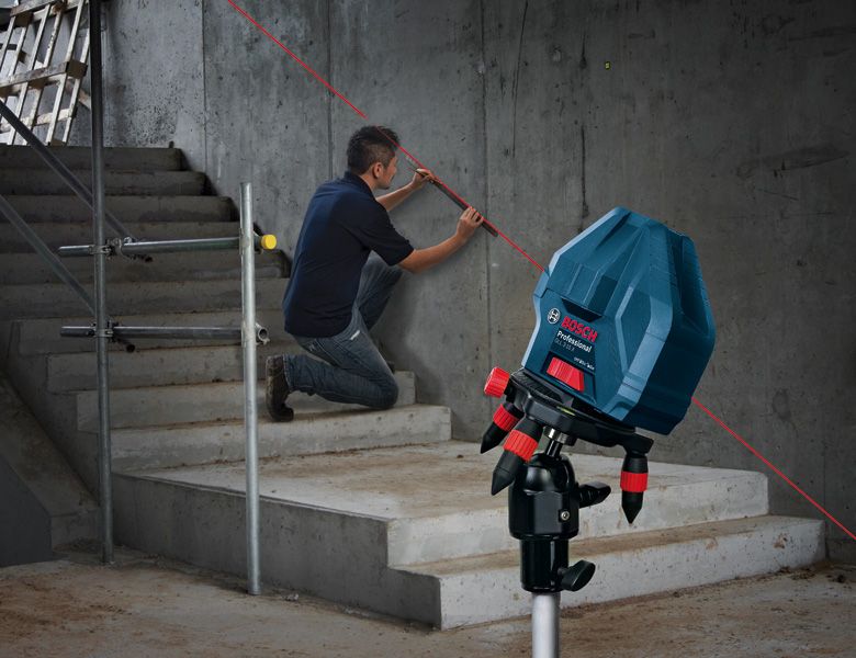 Person kneeling beside stairs uses a laser leveling tool projecting a red line on a concrete wall.