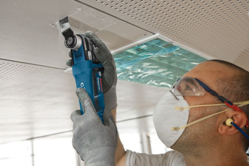 GOP 30-28 A person wearing safety equipment cuts a ceiling panel with a multi-cutter tool.