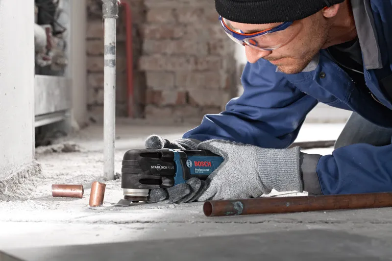 GOP 30-28 A person wearing safety equipment uses a multi-cutter to trim copper pipes on a dusty floor.