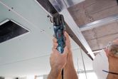 A person wearing safety equipment cuts a ceiling panel with a multi-cutter near metal framing.