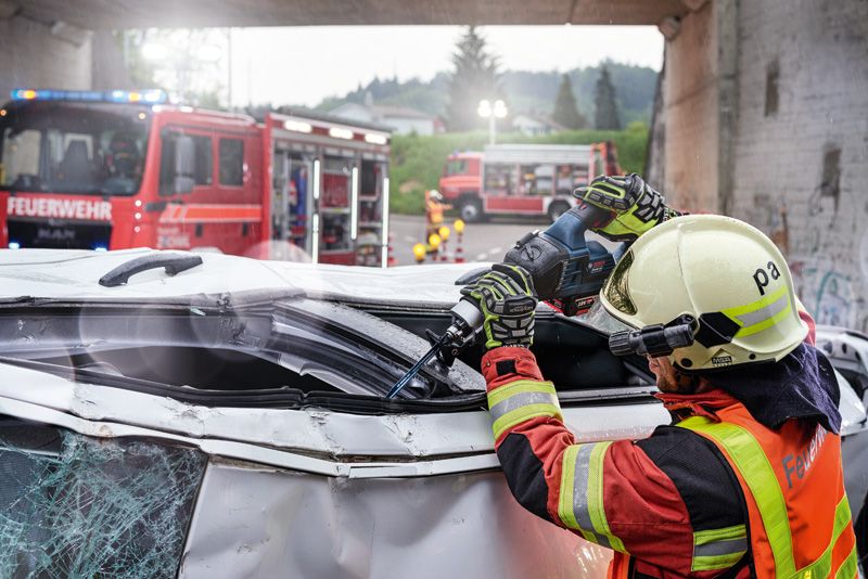 A person wearing safety equipment uses a power tool to cut open a damaged vehicle.