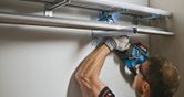 Person wearing safety equipment uses a power drill to mount metal pipes on a wall.