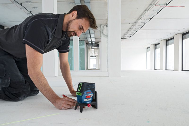 A person marks a line on the floor using a laser leveling tool in a large empty room.