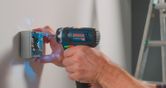 A person uses a cordless drill driver to fasten screws into an electrical box on a wall.