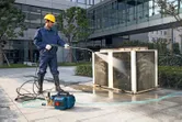 A person wearing safety equipment uses a high-pressure washer to clean an outdoor unit.