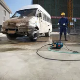 A person wearing safety equipment uses a high-pressure washer to clean a dirty van.