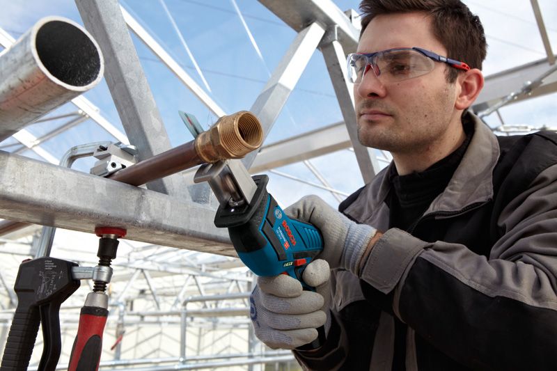 A person wearing safety equipment uses a cordless reciprocating saw to cut a metal pipe.