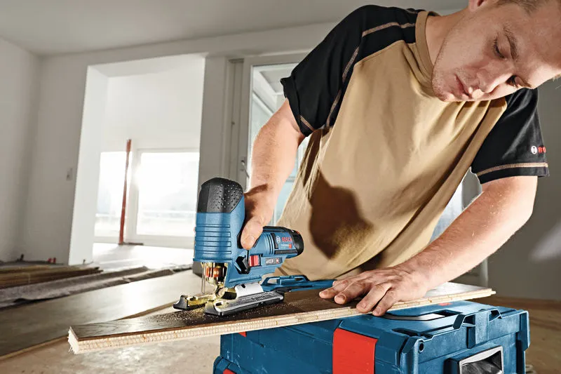 A person cuts a wooden board with a cordless jigsaw on a toolbox.
