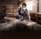 A person wearing safety equipment planes a wooden beam, creating wood shavings.