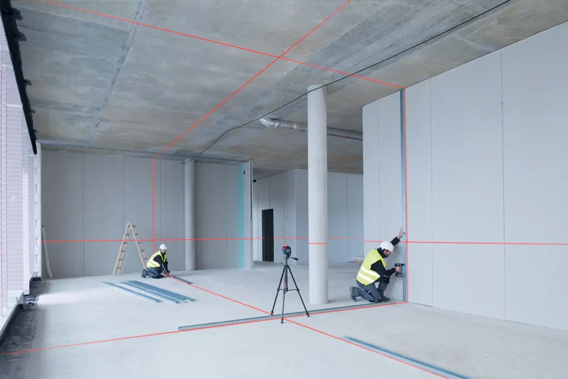 GLL 3-80 Two persons wearing safety equipment align drywall using a laser leveling tool in a building.