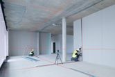 Two persons wearing safety equipment align drywall using a laser leveling tool in a building.