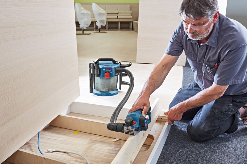 A person wearing safety equipment uses a cordless planer on wooden boards indoors.