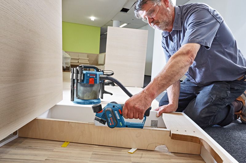A person wearing safety equipment uses a cordless planer to smooth wooden panels.