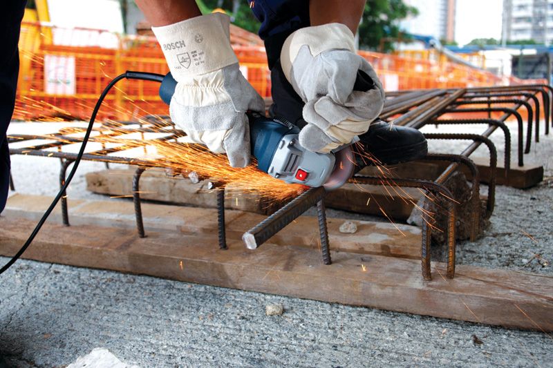 A person wearing safety equipment uses an angle grinder to cut rebar on a construction site.