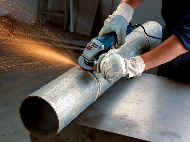 A person wearing safety equipment grinds a large metal pipe with an angle grinder.