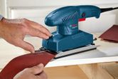 A person fits sandpaper onto an orbital sander placed on a wooden surface.