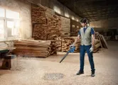 A person wearing safety equipment uses a blower to clear sawdust in a woodworking shop.