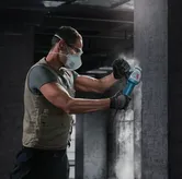 A person wearing safety equipment uses a power grinder on a concrete pillar.