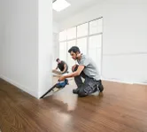 A person cuts flooring near a wall with a cordless saw while another installs floor panels.