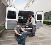 Two workers near an open van organize tools and use a cordless vacuum cleaner.