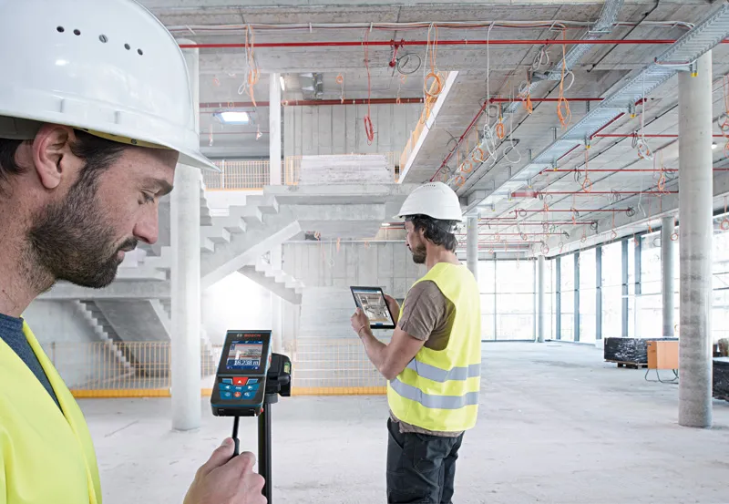 Two people wearing safety equipment use a laser measure and tablet inside a large construction site.