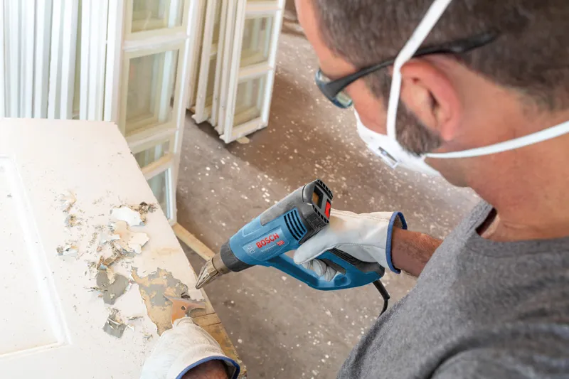 GHG 20-63 A person wearing safety equipment uses a heat gun to strip paint from a door.