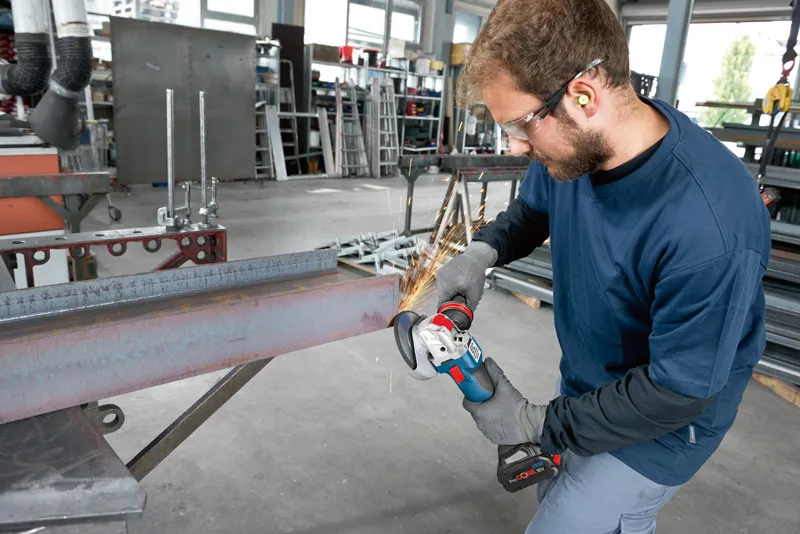 GWX 18V-10 C A person wearing safety equipment grinds a steel beam with a cordless angle grinder.
