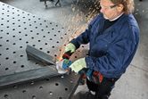 A person wearing safety equipment grinds a metal beam with an angle grinder, creating sparks.